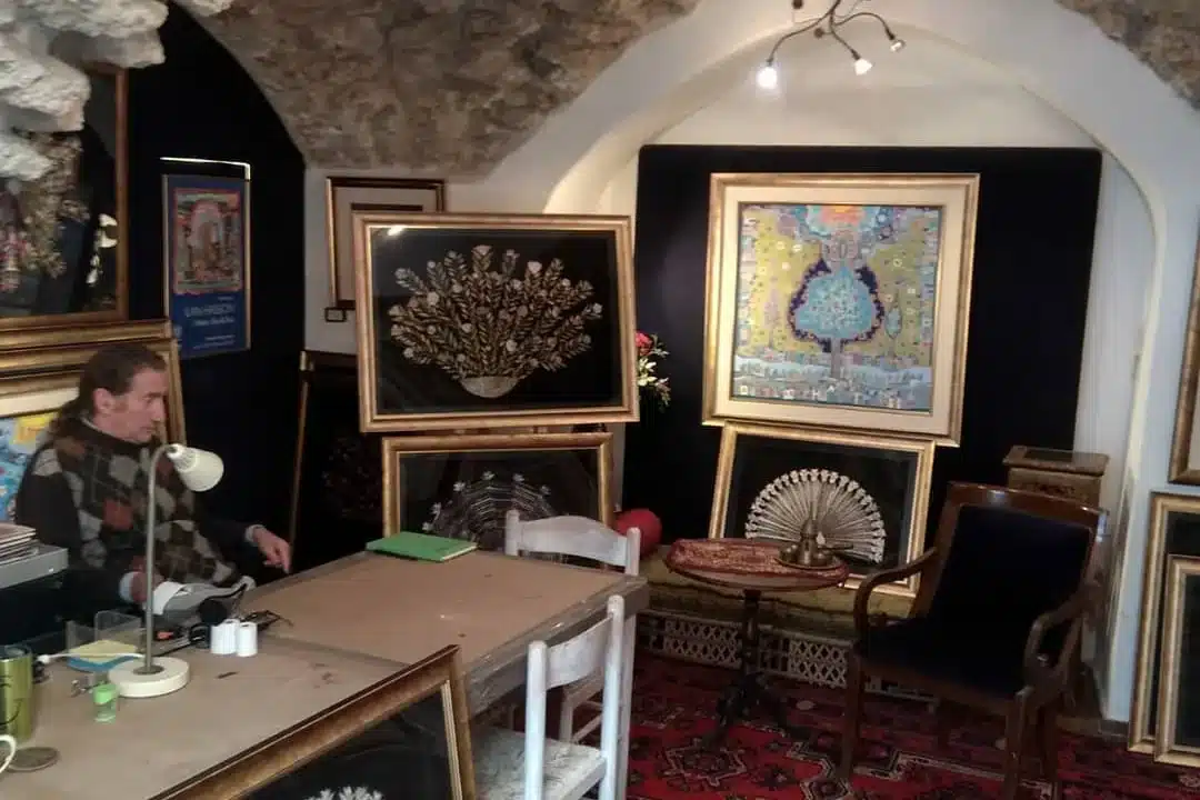 ilan hasson surrounded by artwork Ilan Hasson in his studio, sitting at a desk. Around him many paintings, and some chairs.
